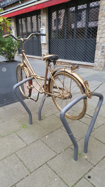 Auf dem Photo ist ein altes Damenfahrrad zu sehen. Es wurde mit goldener Farbe besprüht und dann mit Kabelbinder quer über 2 Fahrradbügel montiert, so dass man diese nicht mehr nutzen kann, um ein Fahrrad anzuschließen.