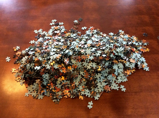 Auf einer braunen Tischplatte liegt ein Haufen ungeordneter Puzzleteile.