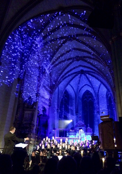 Dirigent rechts mit Chor links  in einer Kirche, deren Kuppel und Wände blau beleuchtet sind, sodass der Eindruck eines Himmels entsteht.