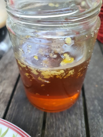 Nahaufnahme eines Glases. Im Glas ist eine goldene Flüssigkeit mit kleinen, gelben Partikeln. Honig mit Resten der Wabe direkt nach dem Schleudern.