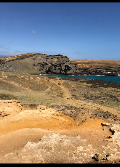 Karge Landschaft mit orange-staubigen Pfaden. Im Hintergrund eine schiefe Kraterkante eines erloschenen Vulkans, der ins Meer ragt. Blaugrauer Himmel