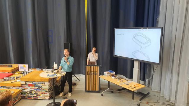 Two guys in front of a large curtain. On the right side is a screen for presentation, on the left a table with the machine