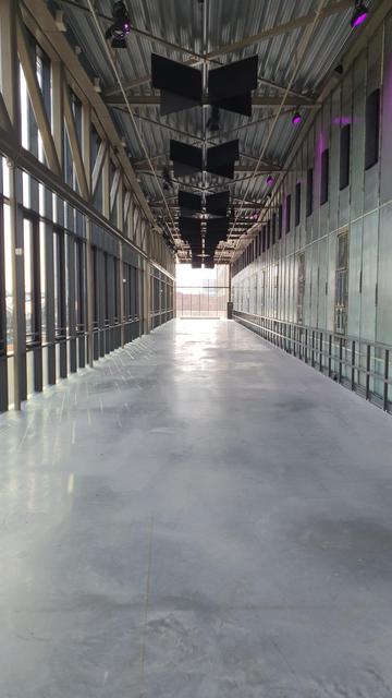 Ein langer Flur in einem Industriegebäude. Am Ende ist ein grosses quadratisches Fenster. Nach links bilden viele schmale Glasscheiben einen seitlichen Abschluss, nach rechts ist der Flur durch Wandelemente begrenzt.