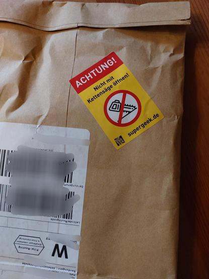 Win brauner Papierumschlag mit einem Aufkleber. Text: Achtung - Nicht mit Kettensäge öffnen
Darunter ein Symbol einer durchgestrichenen Kettensäge und die URL supergeek.com