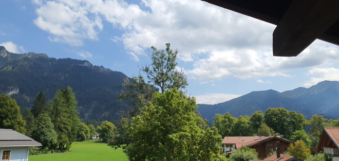 Blick von einem Balkon in die Natur. Am unteren Bildrand sind einige Hausdächer zu sehen. Im Hintergrund sind 2 Bergketten zu sehen.